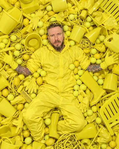 Overhead view of a bearded man lying on his back in a pile of acid-yellow objects, arms spread, looking up