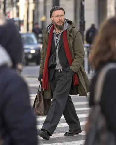 A bearded man in a layered olive parka strides across a crosswalk between two blurred pedestrians in the foreground