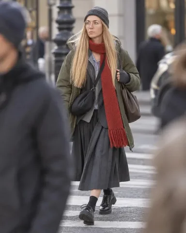 A woman with long blond hair, and glasses in a beanie and red scarf waits at a crossing, city street behind her.