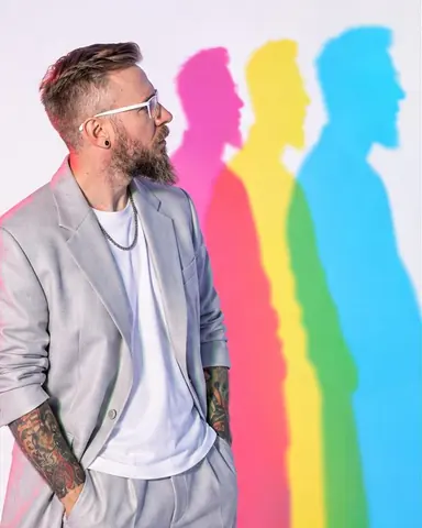 Waist-up profile of a bearded man in white-framed glasses turned to study his own colored shadows with curiosity