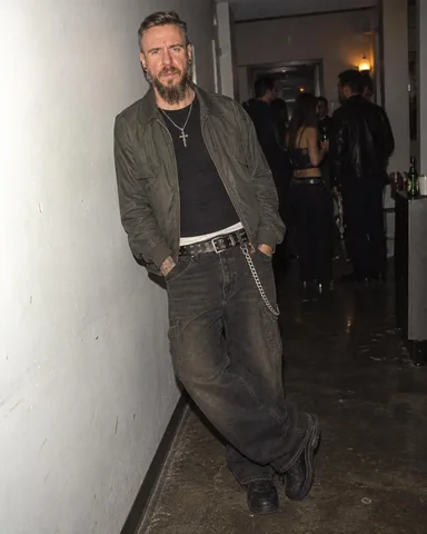 A bearded man with light brown and grey hair leans against a white hallway wall, wearing a dark jacket, black shirt, and chain belt.