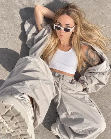 Top-down shot of a woman with long blond hair lying on sunlit concrete in white sunglasses and pale gray parachute trousers