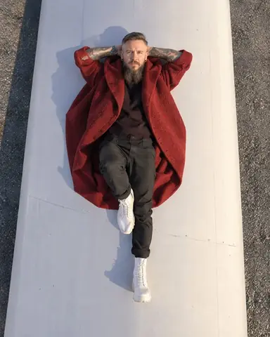 Straight-down drone view of a man in a deep red coat lying on a giant white turbine blade, looking up