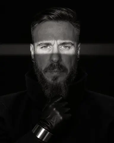 Black-and-white close portrait of a bearded man, a narrow band of hard light crossing his eyes, the rest in black