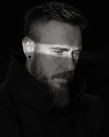 Three-quarter view of a bearded man looking slightly down, a blade of light slicing across his eyes and nose bridge