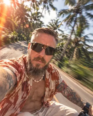 Arm's-length selfie of a bearded man on a scooter in Bali, palm trees smearing behind and an orange sun leak