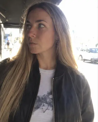 A woman with long blond hair stands under an awning looking aside, the sunlit street blown white with a cyan edge