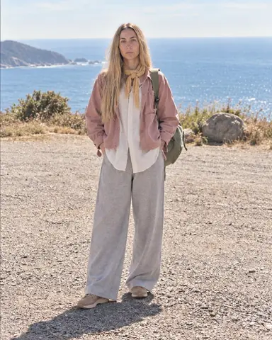 Full length of a woman with long blond hair standing square in a gravel lay-by, her long ecru shirt moving in the breeze