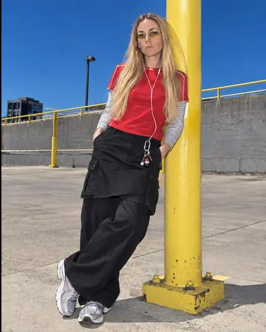 A woman with long blond hair leans on a yellow steel post, hands in pockets, earbud cable on her chest, sky dark cobalt