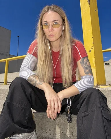 A woman with long blond hair sits on concrete steps, arms resting on knees, in a red shirt and black cargo skirt, yellow rail and post nearby.