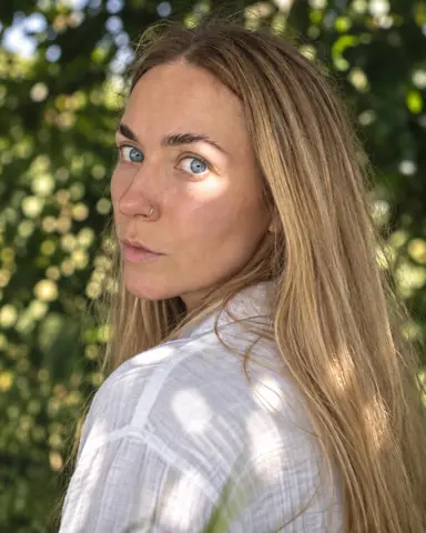 A woman with long blond hair looks back over her shoulder, a round sun spot across one eye and cheek, the rest in shade