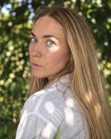 A woman with long blond hair looks back over her shoulder, a round sun spot across one eye and cheek, the rest in shade