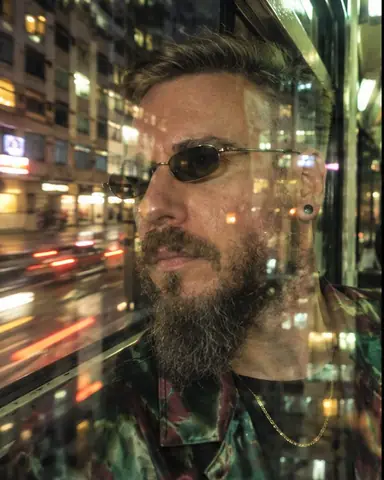 A bearded man's face against a tram window, building reflections over his cheek, the night street smeared outside