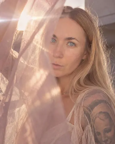 A woman with long blond hair looks through a veil of sheer pink fabric held near the lens, sun flare crossing the frame