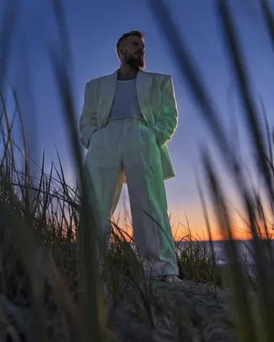 Ground-level view through dune grass of a bearded man in a white linen suit against a deep blue evening sky