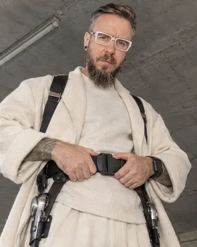 A bearded man in a cream cashmere coat adjusts the hip belt of a carbon exoskeleton, looking down at the camera