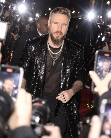 A bearded man in a black sequin coat walks into a storm of flashes, face sharp, five phones in the foreground