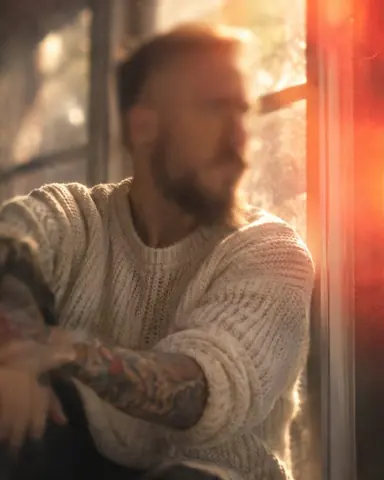 A man in a cream knit stands by a window, a sharp diagonal through his shoulder and a red leak at the right