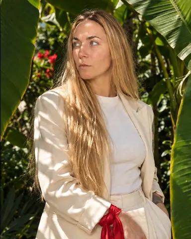Waist-up, a woman with long blond hair between two giant banana leaves, head turned, leaf shadows striping her cream suit