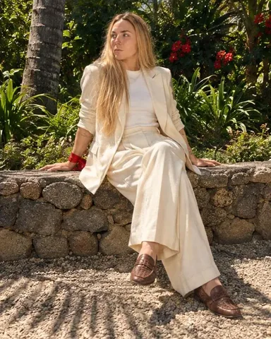 Waist-up, a woman with long blond hair between two giant banana leaves, head turned, leaf shadows striping her cream suit