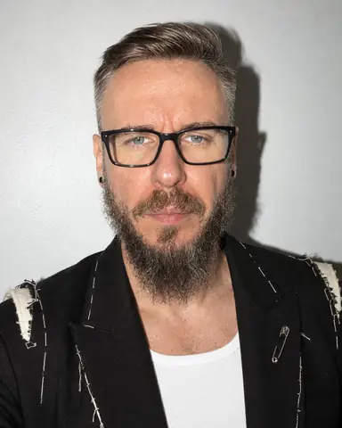 Tight portrait of a bearded man in black glasses against a white wall, eyes to the lens, a hard flash shadow behind