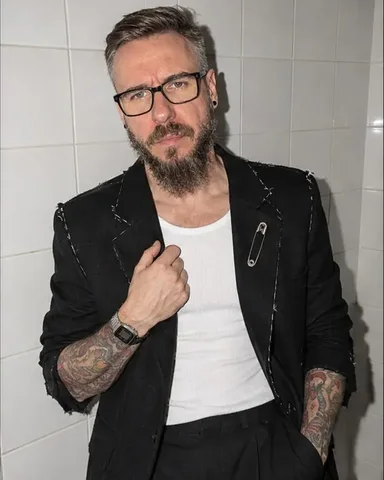 A man in a raw-edged black blazer leans on white bathroom tiles, one hand at the lapel, lit by a bare flash