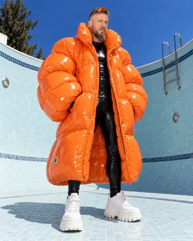 A bearded man in a huge glossy orange inflatable coat stands in the deep end of an empty pool against pale tiles