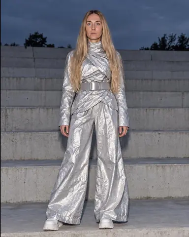 A woman with long blond hair in a crinkled silver suit stands on a concrete step, terraces close behind, in flat flash