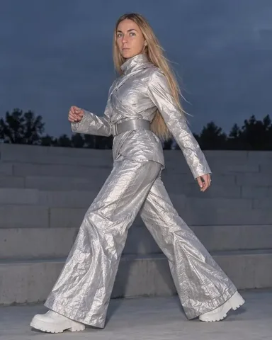 A woman with long blond hair in a crinkled silver suit stands on a concrete step, terraces close behind, in flat flash