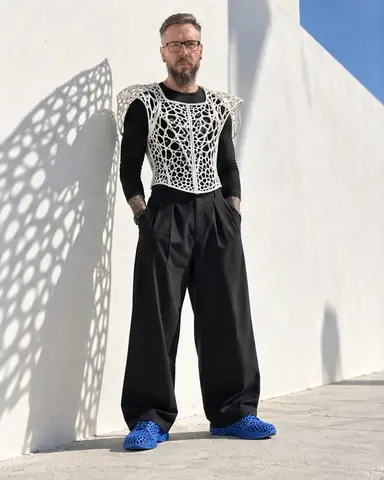 A bearded man in a white lattice vest stands against a white wall, its shadow projected large beside him