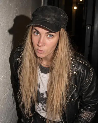A woman with long blond hair under a waxed cap leans on a scuffed corridor wall, her head's hard shadow thrown sideways