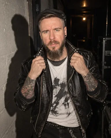 A bearded man with light brown hair leans against a wall in a narrow backstage corridor, hands in pockets.