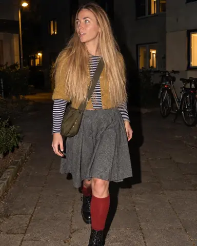 A woman with long blond hair walks past the camera through a dark courtyard, glancing sideways, her flash shadow on the wall