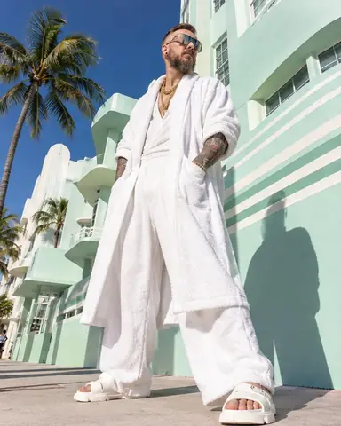 Low-angle shot of a bearded man in a white terry robe-coat against a mint art deco facade and deep blue sky