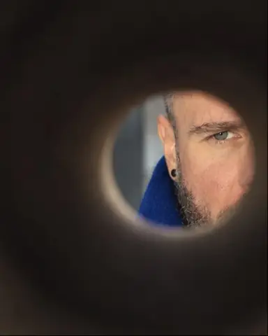 Through a cardboard tube, a small circle at lower right reveals a man's eye and cobalt collar, the rest soft black