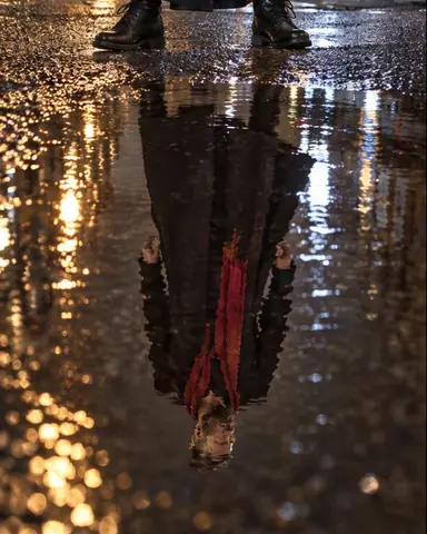 Upside-down reflection of a man in a long black coat in a rippled night puddle, his real boots at the top edge