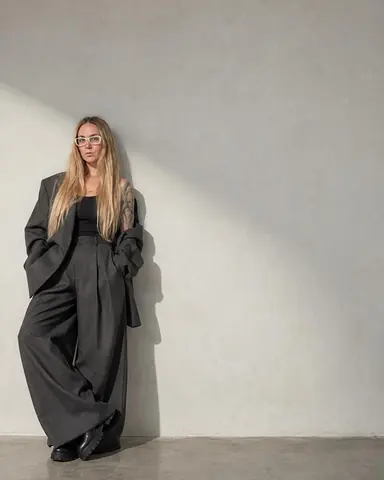 A woman with long blond hair in white glasses leans small at the lower left of a vast pale wall, oversized gray suit