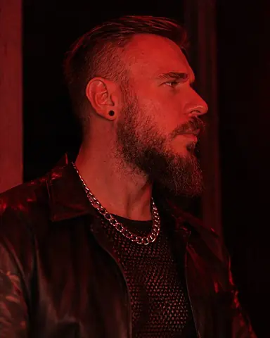 Profile of a bearded man against a black doorway, red light tracing his face, silver chains glinting at the neck