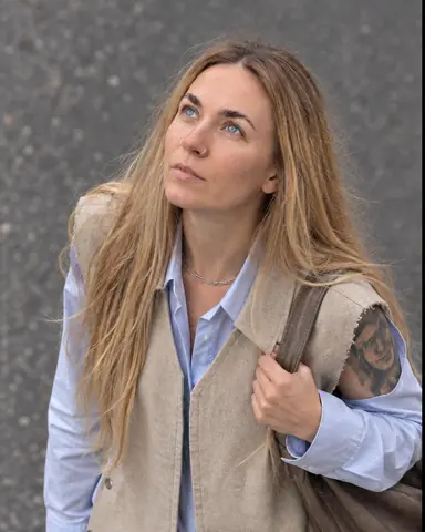 Waist-up from a window above, a woman with long blond hair walks with her face tilted up to the sky, hand on her bag strap