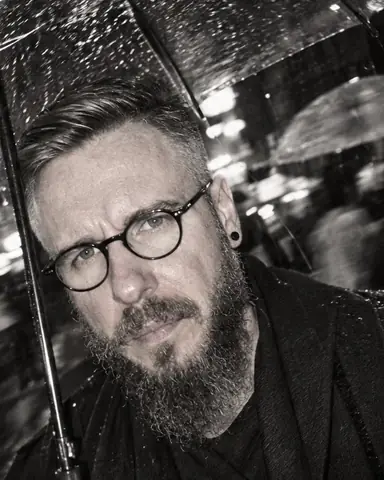 Tilted close-up of a bearded man lit by hard flash, raindrops frozen midair, blurred umbrellas behind, black and white