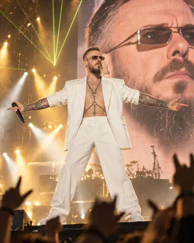 Wide view from the pit of a bearded man at the stage edge, arms spread, beams above and his face on a giant LED screen
