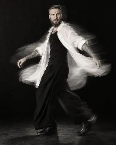 Black-and-white full-length of a bearded man spinning, his white shirt flaring into a curved trail, face frontal