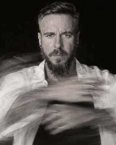 Black-and-white full-length of a bearded man spinning, his white shirt flaring into a curved trail, face frontal