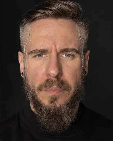 A bearded man raises his hands before his face, silver traces across the knuckles, eyes between his fingers