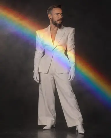 Full-length bearded man in a white hourglass suit, a diagonal rainbow band across his body, the rest dark