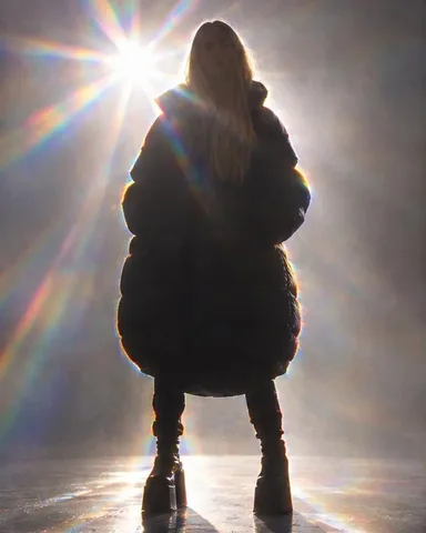 A woman with long blond hair in a black puffer coat and platform boots, standing against a bright backlight with rainbow flares.