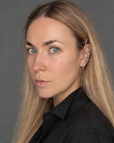 A woman with long blond hair turned three-quarter, eyes to the lens, a sculptural ear-climber sharp against a gray wall