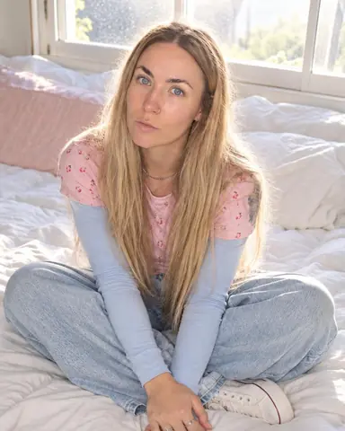 A woman with long blond hair sits cross-legged on a white duvet, looking up, the window behind glowing milky white