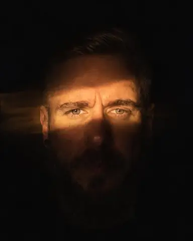 A band of warm flashlight light falls across a bearded man's eyes and nose, the rest of the frame black