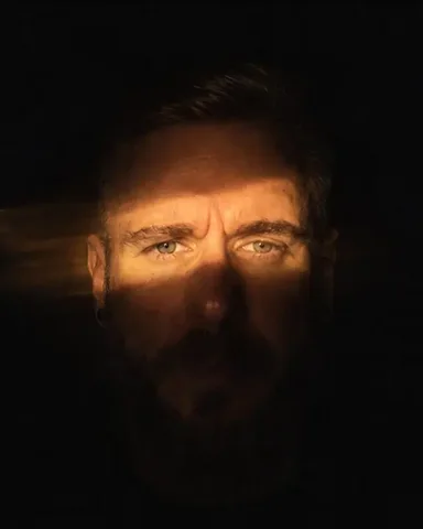 A band of warm flashlight light falls across a bearded man's eyes and nose, the rest of the frame black